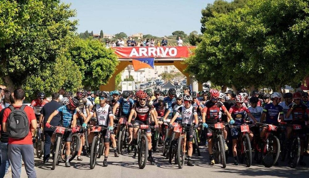 Partenza della Granfondo MTB a Santa Ninfa con decine di ciclisti pronti alla gara sotto lo striscione "Arrivo", tra due ali di alberi e pubblico festante.
