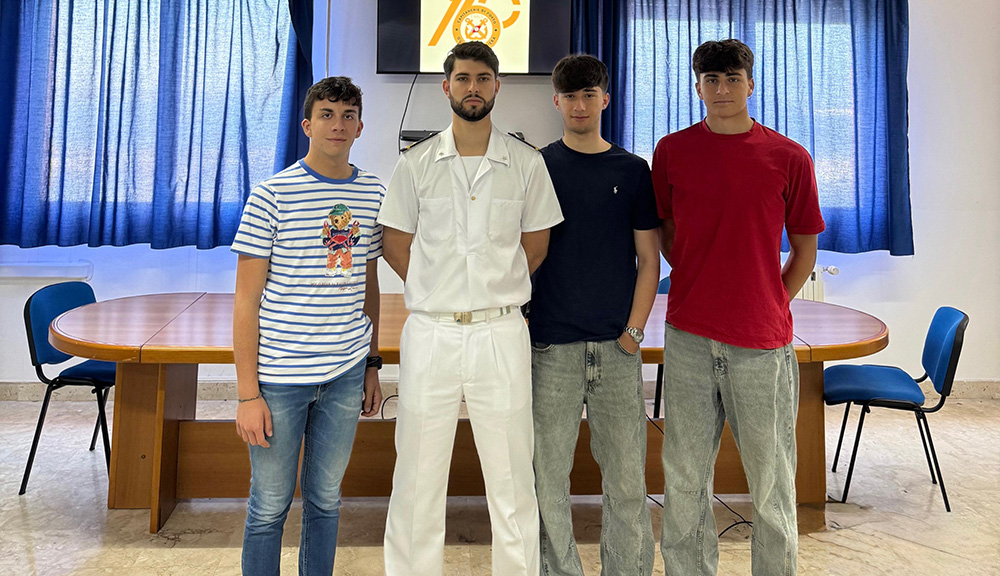 "Quattro studenti dell'Istituto Nautico 'Leonardo da Vinci' di Trapani, uno in uniforme bianca della Guardia Costiera, posano all'interno di una sala riunioni con tavolo ovale e tende blu."