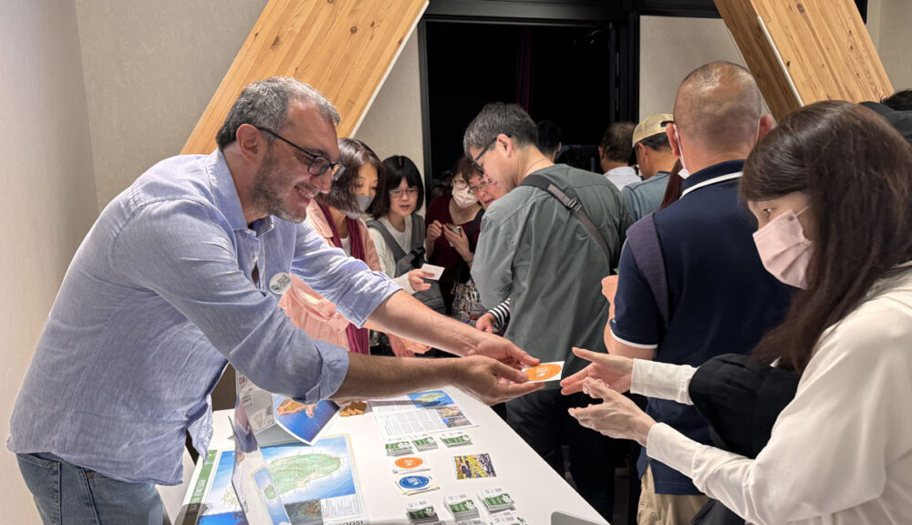 "Uomo con camicia chiara sorride mentre consegna materiale promozionale a visitatori giapponesi presso uno stand informativo, durante un evento culturale o turistico internazionale."