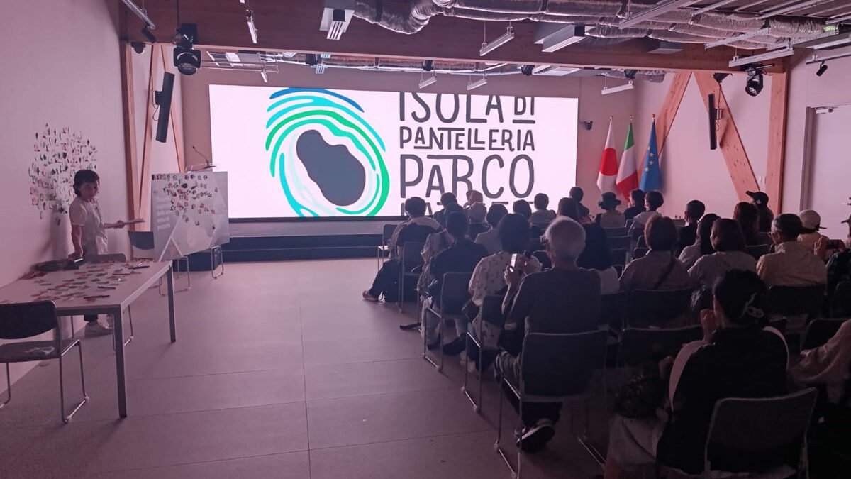 "Presentazione del Parco Nazionale Isola di Pantelleria a un pubblico giapponese in sala conferenze, con maxi-schermo, bandiere italiana, giapponese ed europea, durante un evento promozionale internazionale."
