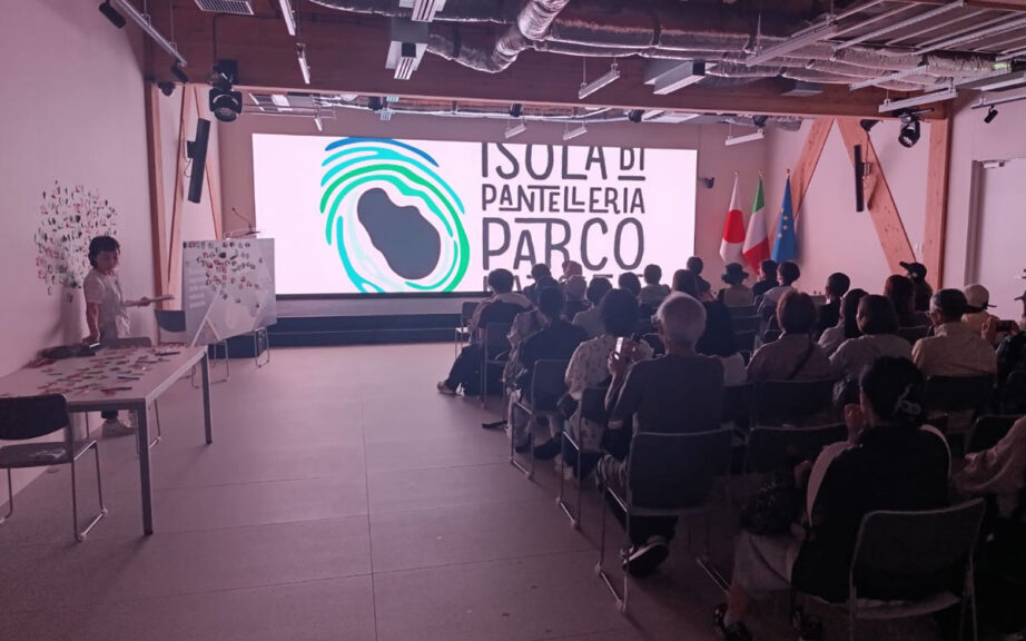 "Presentazione del Parco Nazionale Isola di Pantelleria a un pubblico giapponese in sala conferenze, con maxi-schermo, bandiere italiana, giapponese ed europea, durante un evento promozionale internazionale."