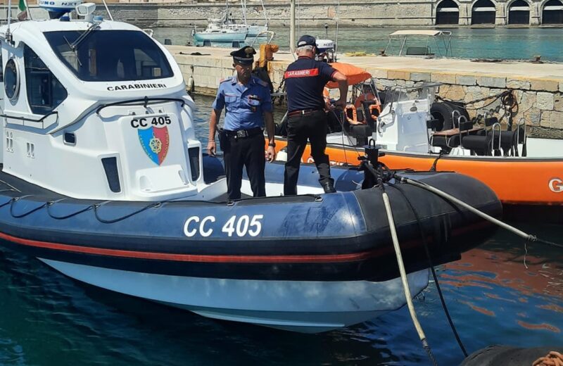 Motovedetta CC 405 dei Carabinieri ormeggiata nel porto di Favignana, con due militari a bordo in divisa operativa. Sullo sfondo, edifici storici e acqua cristallina dell'Area Marina Protetta delle Isole Egadi.