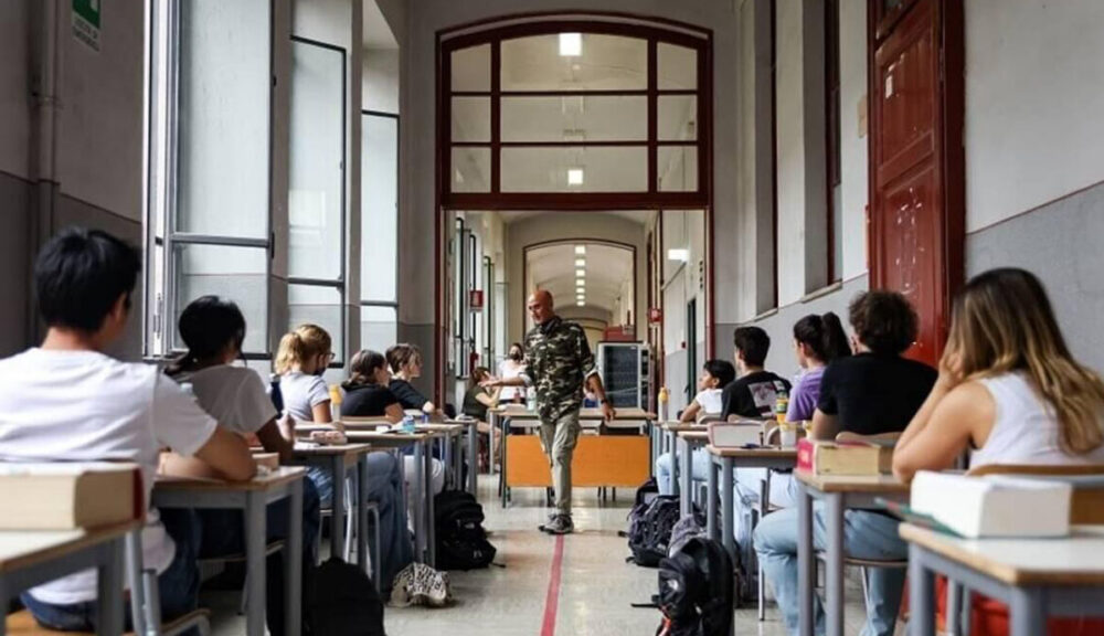 Studenti durante la prima prova dell’esame di maturità, seduti ai banchi nel corridoio di una scuola, sotto lo sguardo del docente sorvegliante.
