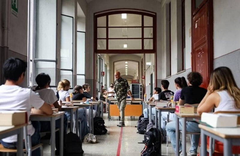 Studenti durante la prima prova dell’esame di maturità, seduti ai banchi nel corridoio di una scuola, sotto lo sguardo del docente sorvegliante.
