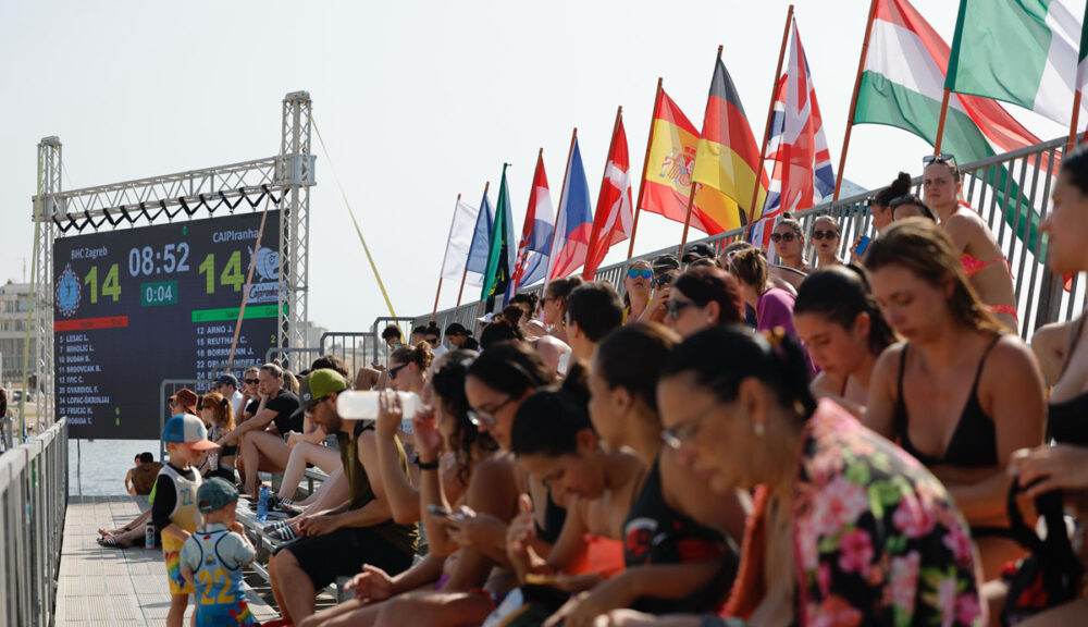 Spettatori sugli spalti durante le EBT Finals 2025 di beach handball a Trapani, con bandiere europee e tabellone del match BHC Zagreb – CAIPiranhas sullo sfondo.