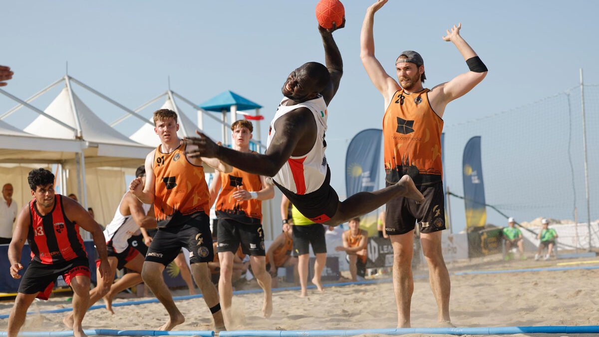 Giocatore in volo durante un'azione spettacolare di beach handball maschile alle EBT Finals 2025 di Trapani, con avversari in difesa sulla sabbia.