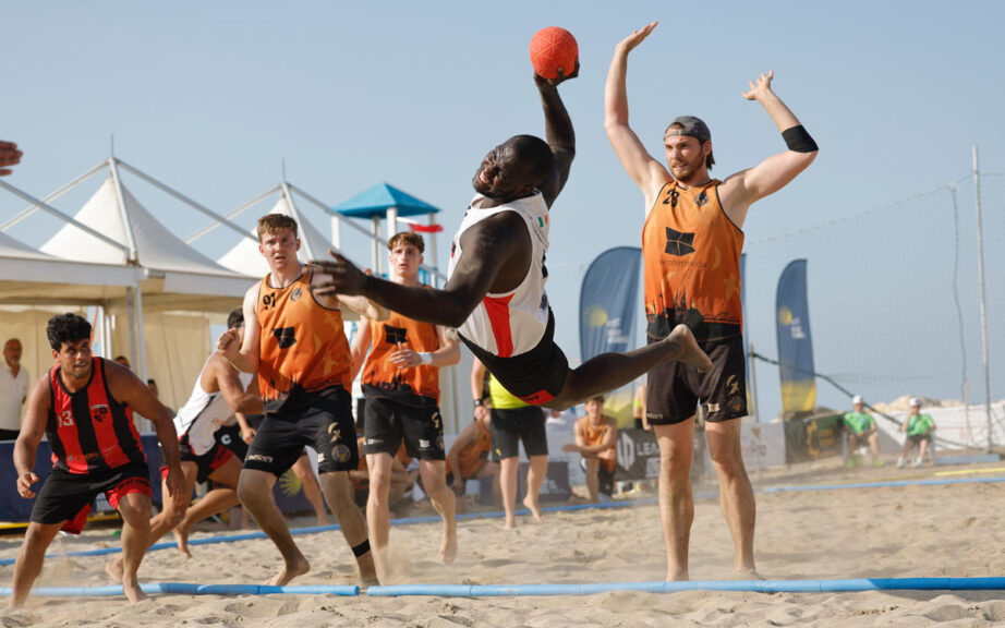 Giocatore in volo durante un'azione spettacolare di beach handball maschile alle EBT Finals 2025 di Trapani, con avversari in difesa sulla sabbia.
