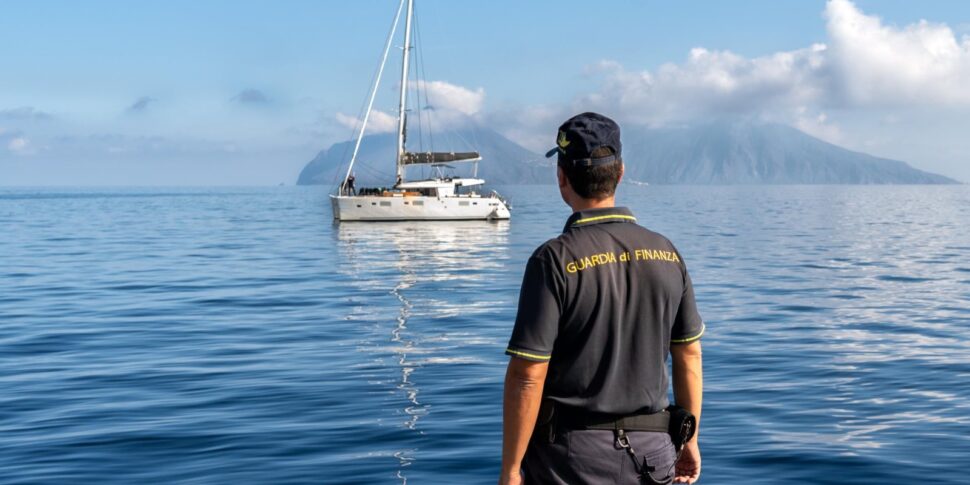 Agente della Guardia di Finanza osserva uno yacht a vela al largo di Milazzo, con le isole Eolie sullo sfondo