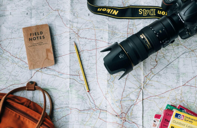 Accessori da viaggio su mappa: fotocamera Nikon, taccuino Field Notes, zaino arancione, matita e cartine geografiche per la pianificazione di un itinerario.