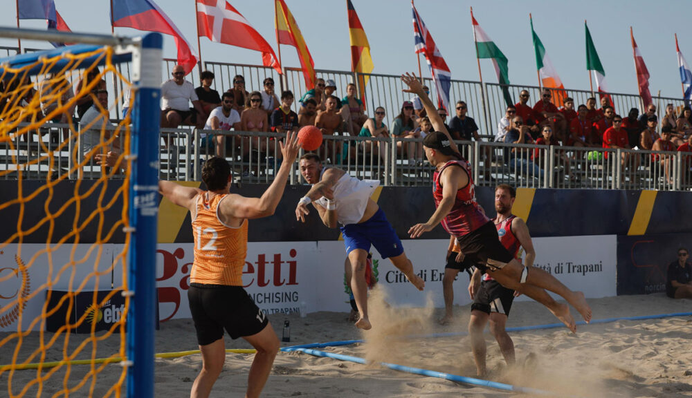 Partita di beach handball alle EBT Finals 2025 a Trapani con atleti in volo sulla sabbia, pubblico sugli spalti e bandiere internazionali sullo sfondo.