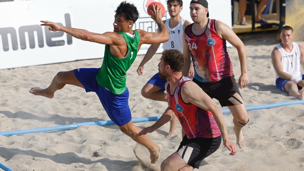 Giocatore di beach handball in volo mentre tenta un tiro, contrastato da due avversari, durante una partita su sabbia. Sullo sfondo altri atleti e spettatori.