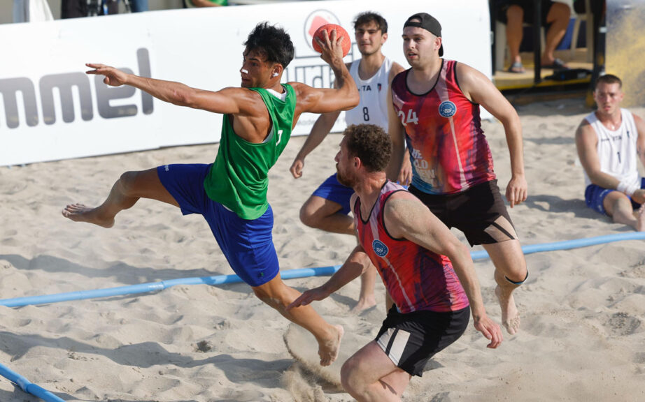 Giocatore di beach handball in volo mentre tenta un tiro, contrastato da due avversari, durante una partita su sabbia. Sullo sfondo altri atleti e spettatori.