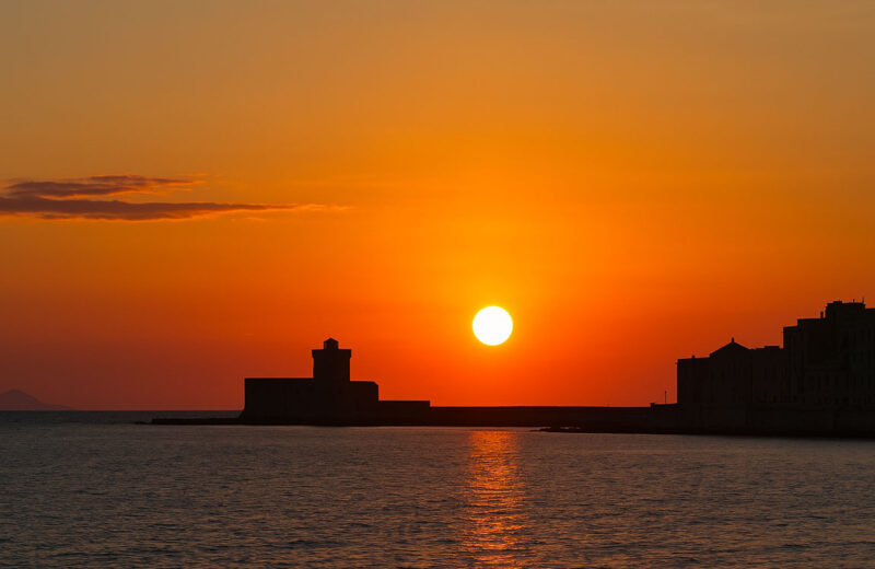 Tramonto estivo sul mare a Trapani durante il solstizio, con il sole basso sull’orizzonte, cielo arancione e silhouette della città costiera.