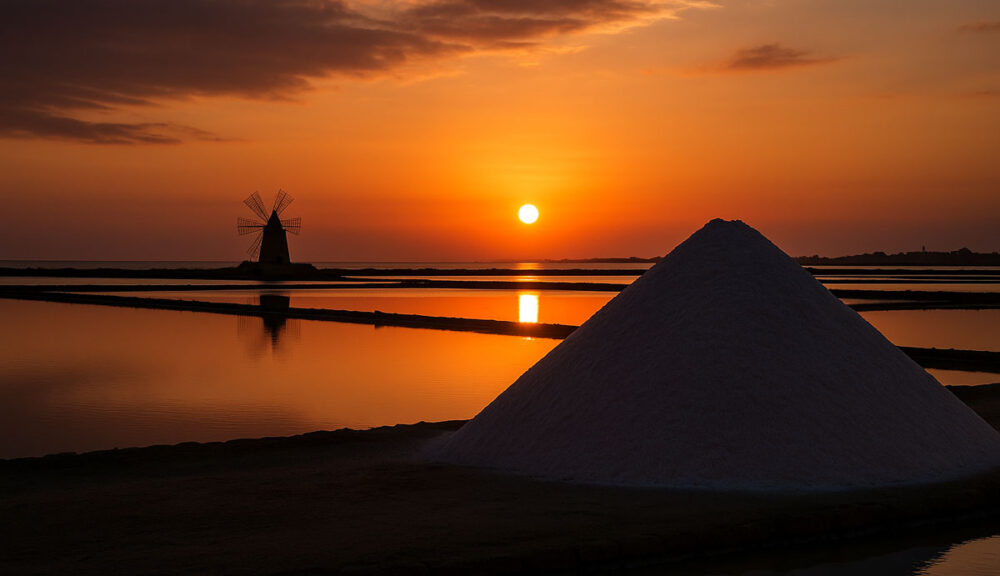 Tramonto sulle saline di Trapani con mulino a vento e cumulo di sale riflessi nell’acqua calma.