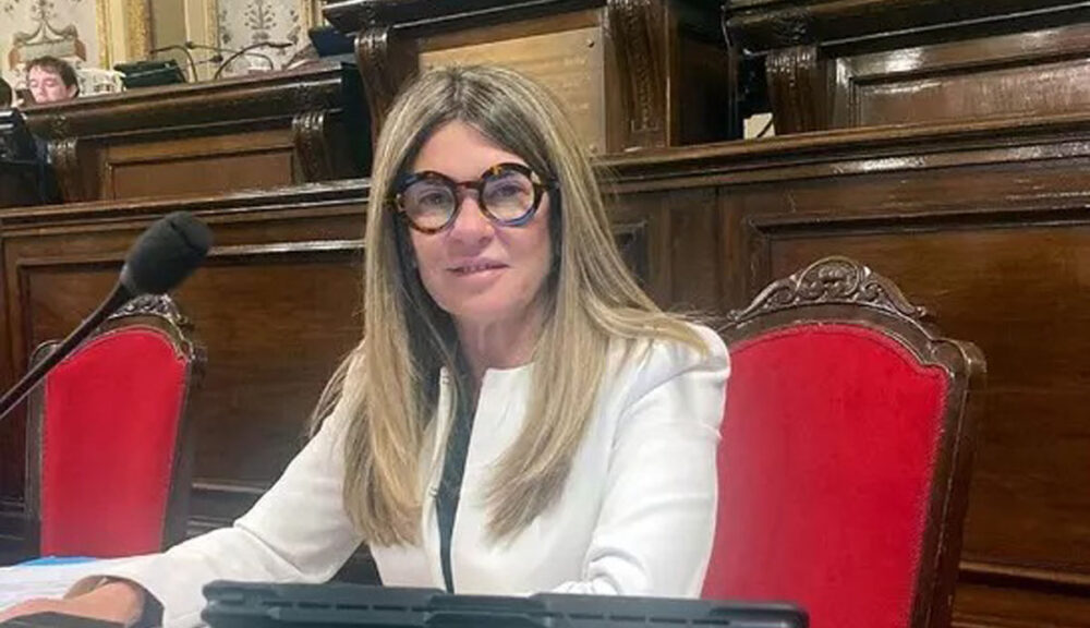 Donna seduta su una poltrona rossa in un’aula istituzionale, con abito bianco e occhiali scuri, davanti a un microfono e a un tablet.