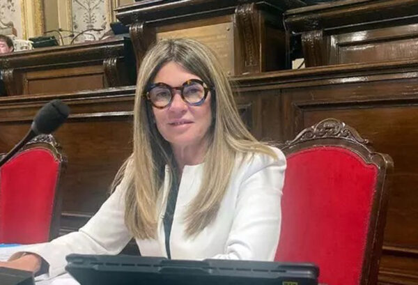Donna seduta su una poltrona rossa in un’aula istituzionale, con abito bianco e occhiali scuri, davanti a un microfono e a un tablet.