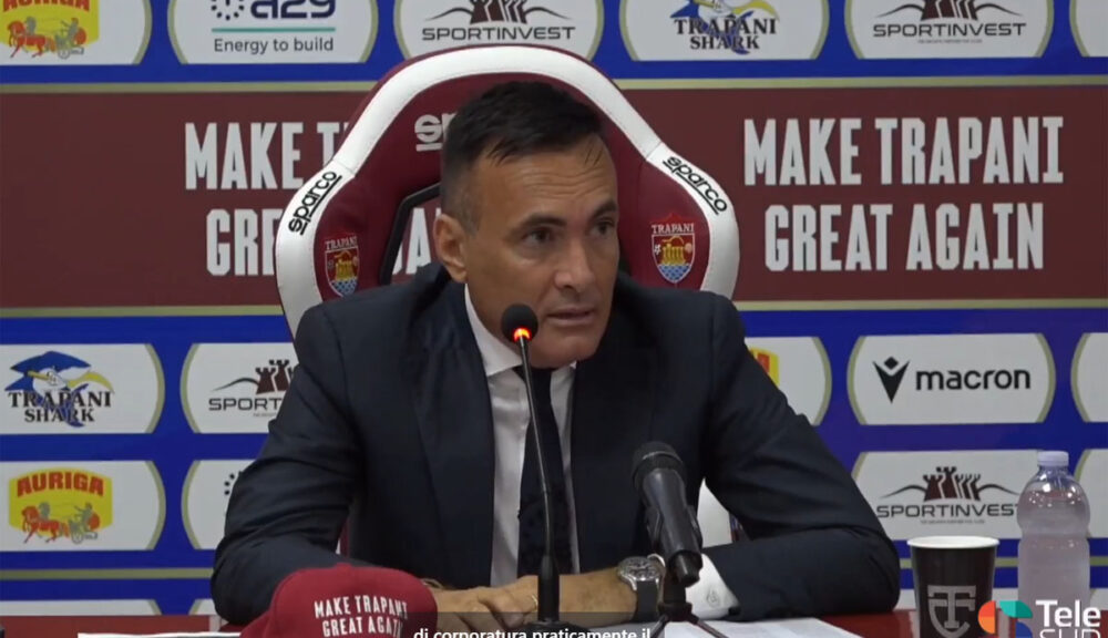 Valerio Antonini durante una conferenza stampa a Trapani, seduto su una sedia Sparco con lo slogan "Make Trapani Great Again" sullo sfondo. Sul tavolo un cappellino con la stessa scritta, microfoni, bicchiere con logo TT e bottiglia d’acqua. Sullo sfondo i loghi di Trapani Shark, SportInvest, Macron, A29 e Auriga. Logo di TeleSud in basso a destra. Chiedi a ChatGPT