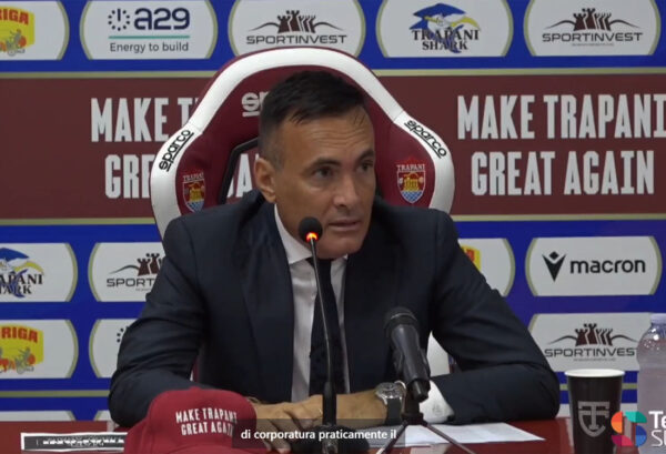 Valerio Antonini durante una conferenza stampa a Trapani, seduto su una sedia Sparco con lo slogan "Make Trapani Great Again" sullo sfondo. Sul tavolo un cappellino con la stessa scritta, microfoni, bicchiere con logo TT e bottiglia d’acqua. Sullo sfondo i loghi di Trapani Shark, SportInvest, Macron, A29 e Auriga. Logo di TeleSud in basso a destra. Chiedi a ChatGPT