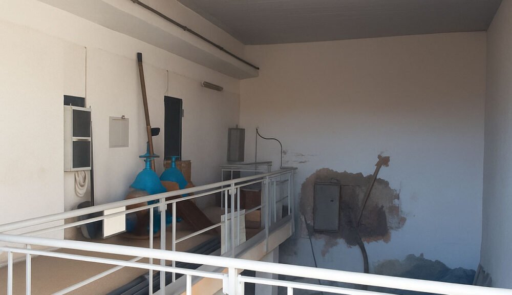 Locale tecnico con tubazioni, valvole blu e quadri elettrici su parete bianca con segni di umidità